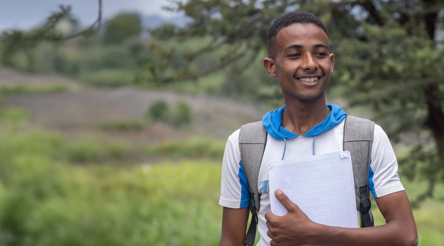 Munadhel, a university student from Yemen, smiles whilst wearing a backpack. He is holding a stack of papers.