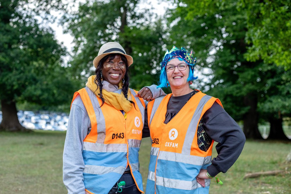 Two Oxfam stewards smiling for the camera as they lean on each other's shoulders in front of the trees at Wilderness Festival