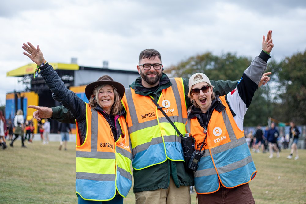 Three smiling Oxfam Festival volunteers with arms raised in sky as they pose in front of the main stage at Leeds Festival