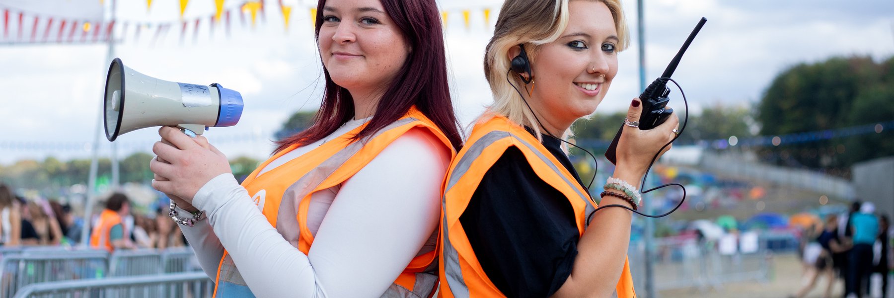 Two Oxfam volunteer Stewards standing back to back as they pose at the entrance to the main arena at Leeds Festival. One steward is holding a megaphone and the other is holding a radio.