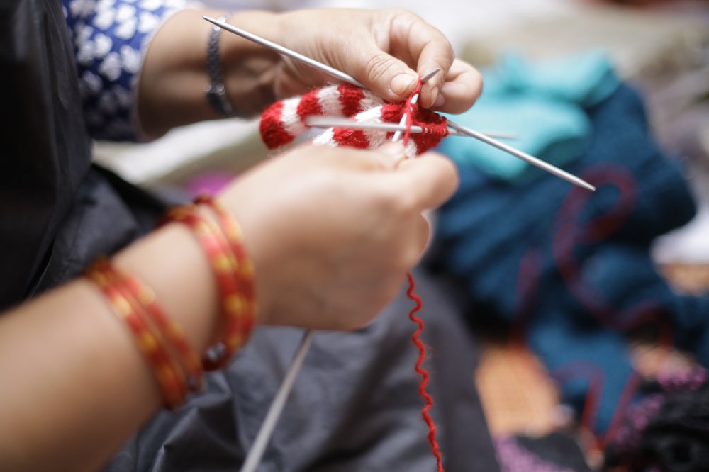 A pair of hands knitting a red and white jumper-shaped Christmas tree decoration