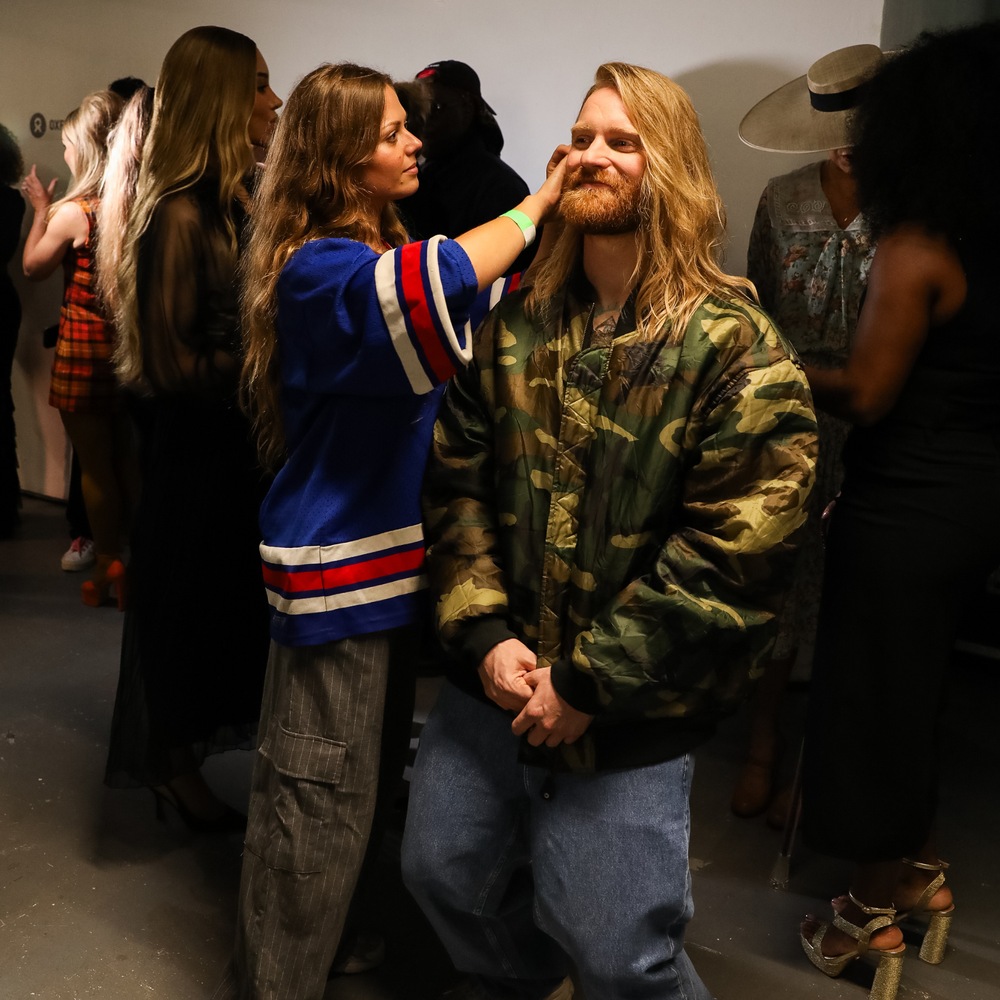 Sam Ryder backstage getting ready for the Oxfam x Vinted Style for Change fashion show. He wears a camo puffa jacket and jeans.