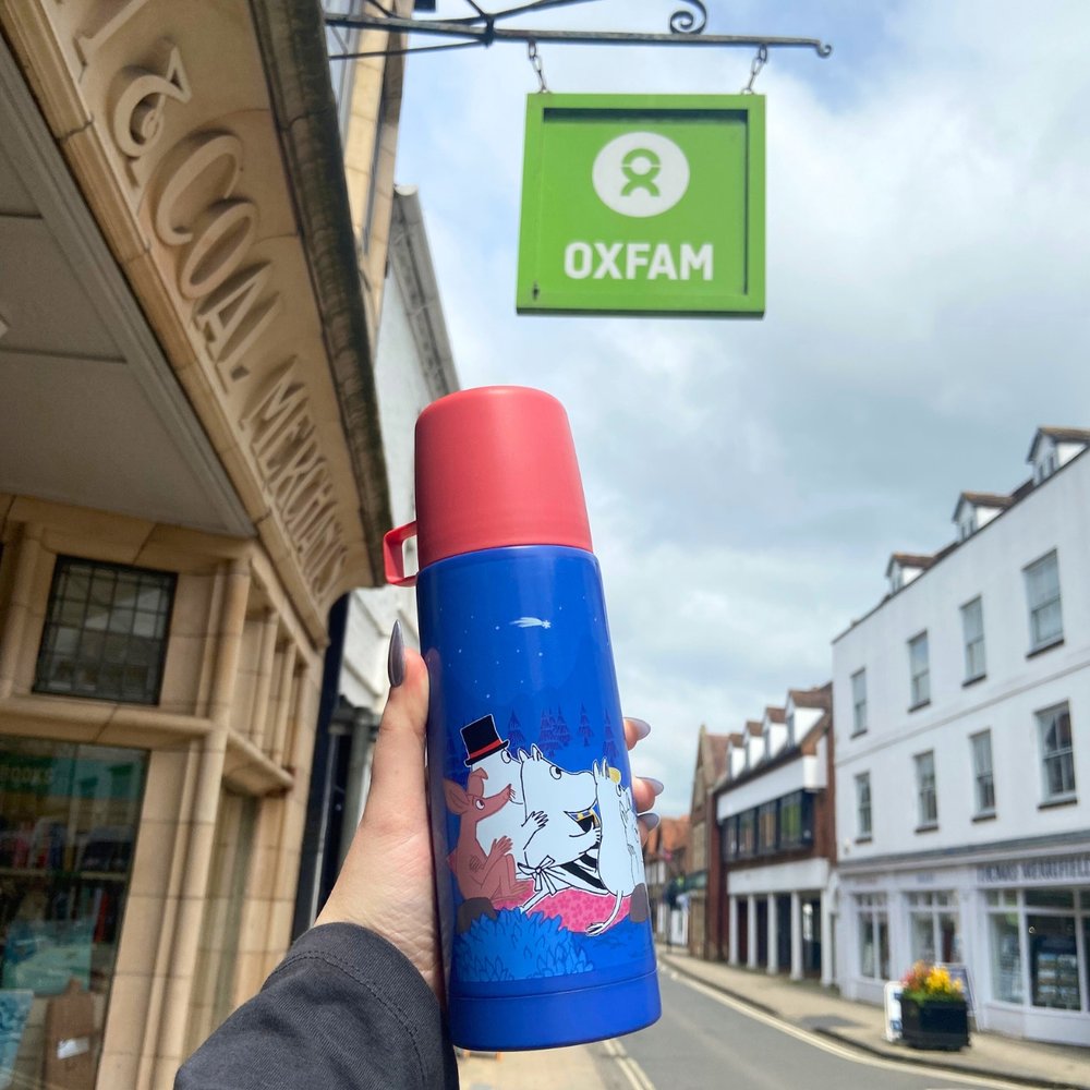 Red and blue thermal flask with Moomin illustrations held up in front of an Oxfam shop