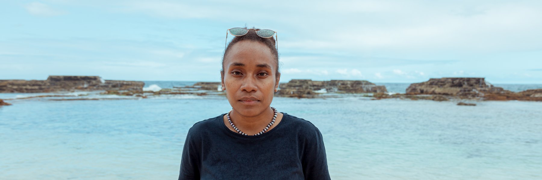 Cynthia looks into the camera. Behind her is the beach and the sea with rocks jutting out of it.