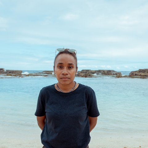 Cynthia looks into the camera. Behind her is the beach and the sea with rocks jutting out of it.