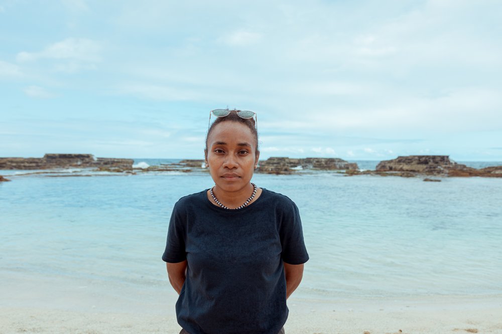 Cynthia looks into the camera. Behind her is the beach and the sea with rocks jutting out of it.