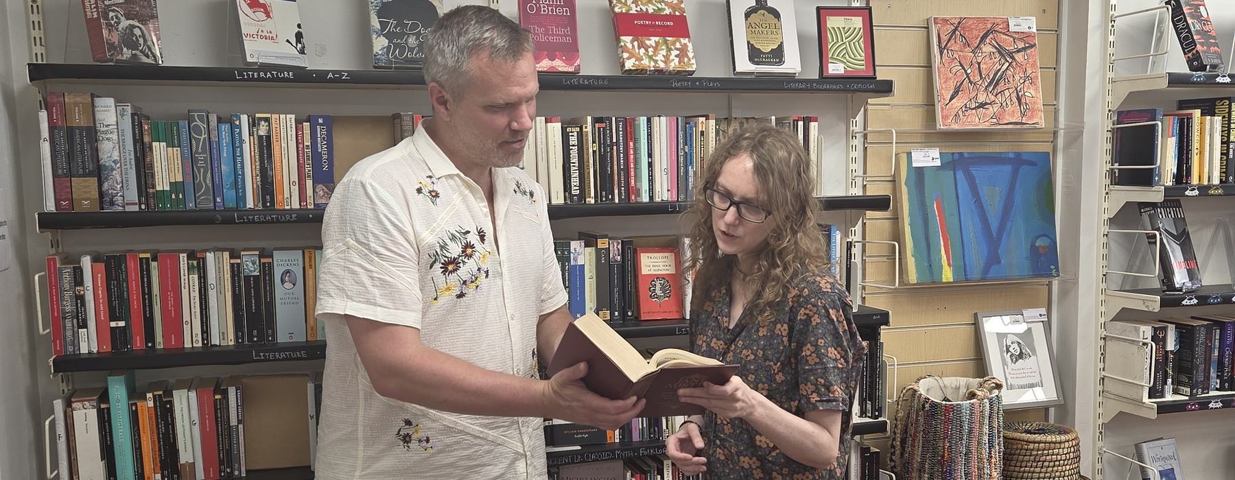 Two people stand in a bookshop browsing shelves, reading from an open book together.
