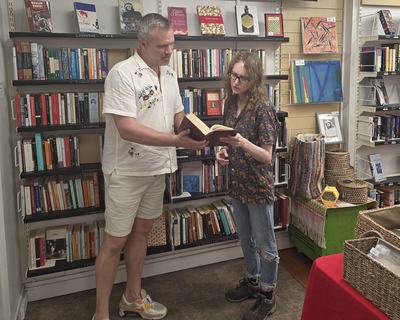 Two people stand in a bookshop browsing shelves, reading from an open book together.
