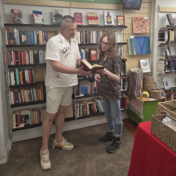 Two people stand in a bookshop browsing shelves, reading from an open book together.