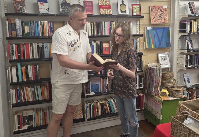 Two people stand in a bookshop browsing shelves, reading from an open book together.