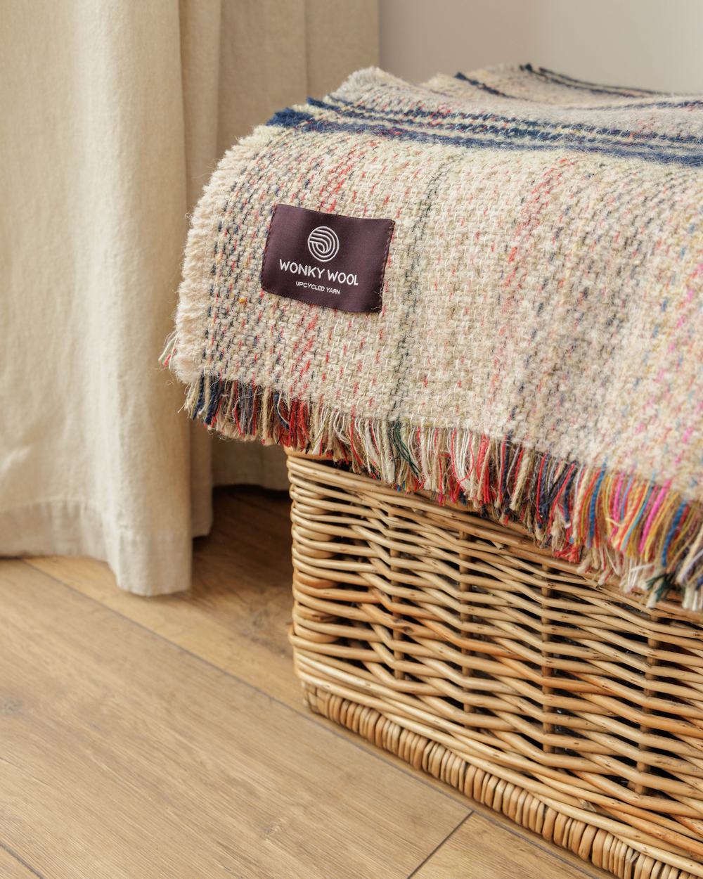 A textured recycled wool throw with multicoloured flecks draped over a wicker basket beside a curtain.