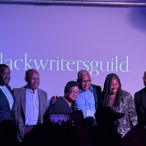 A group of six people stand on stage in front of a screen displaying “The Black Writers Guild,” smiling as the audience takes photos.