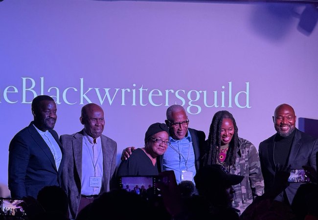 A group of six people stand on stage in front of a screen displaying “The Black Writers Guild,” smiling as the audience takes photos.