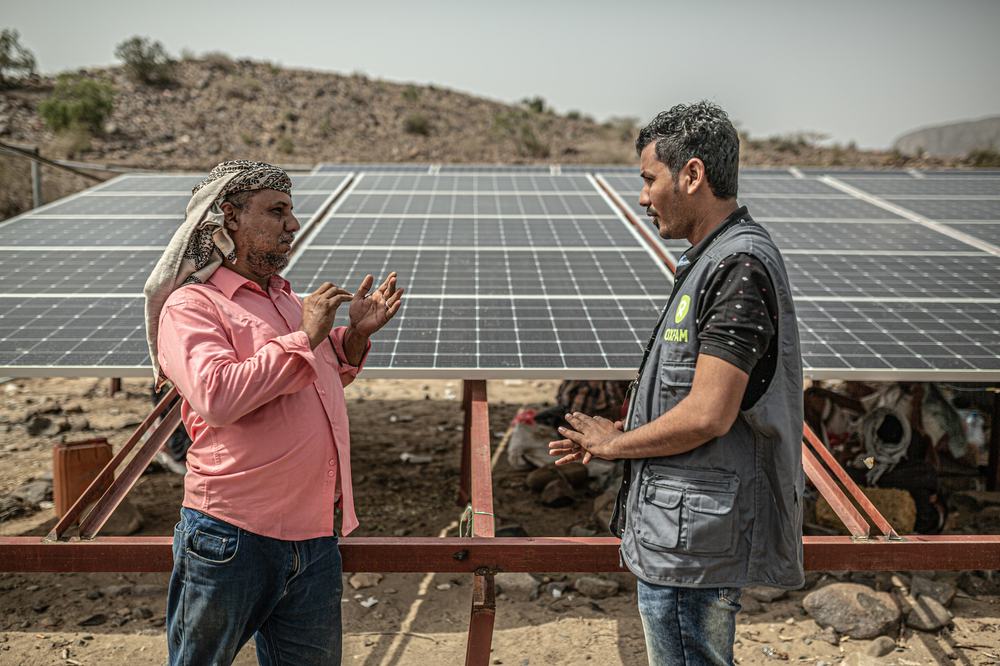 An Oxfam Aid worker and a man with a scarf around his head talk in front of Oxfam solar panels