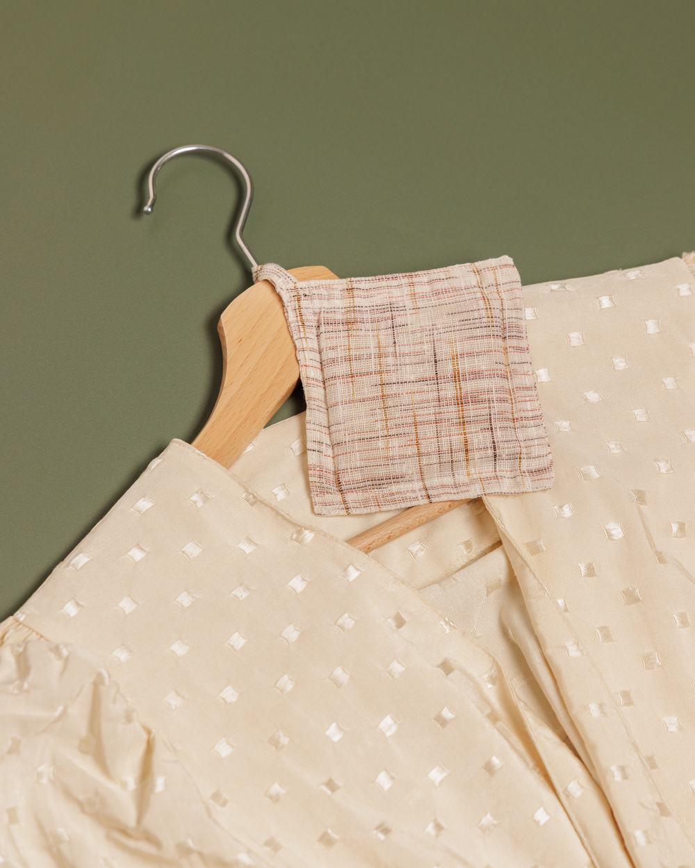 A square gingham cloth scent bag tied to a wooden hanger and cream-coloured blouse, laid flat against a khaki green surface.