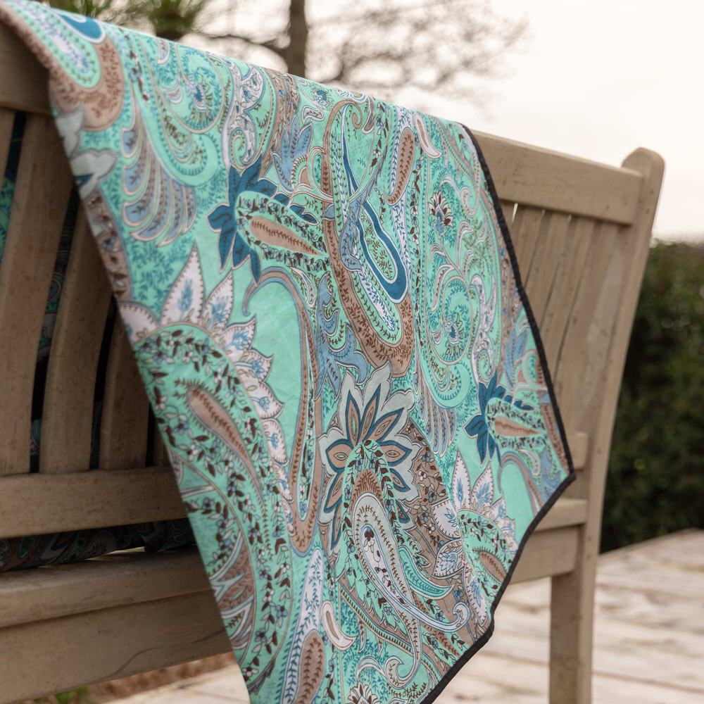 A green paisley print picnic blanket hanging on the back of a wooden bench.