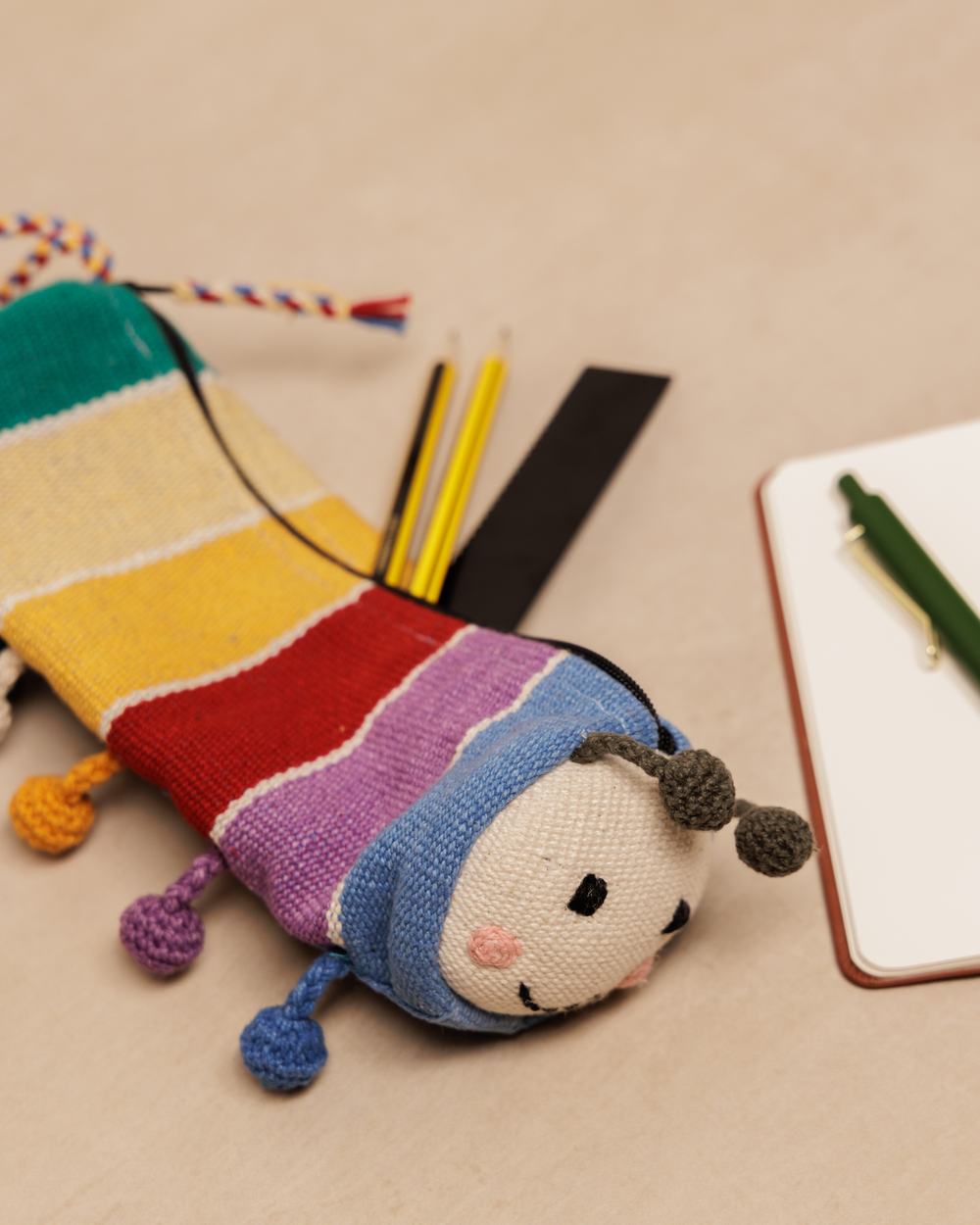 A colourful fabric pencil case shaped like a stripey, smiley caterpillar, laid flat, next to various stationery items such as pencils and a notebook.