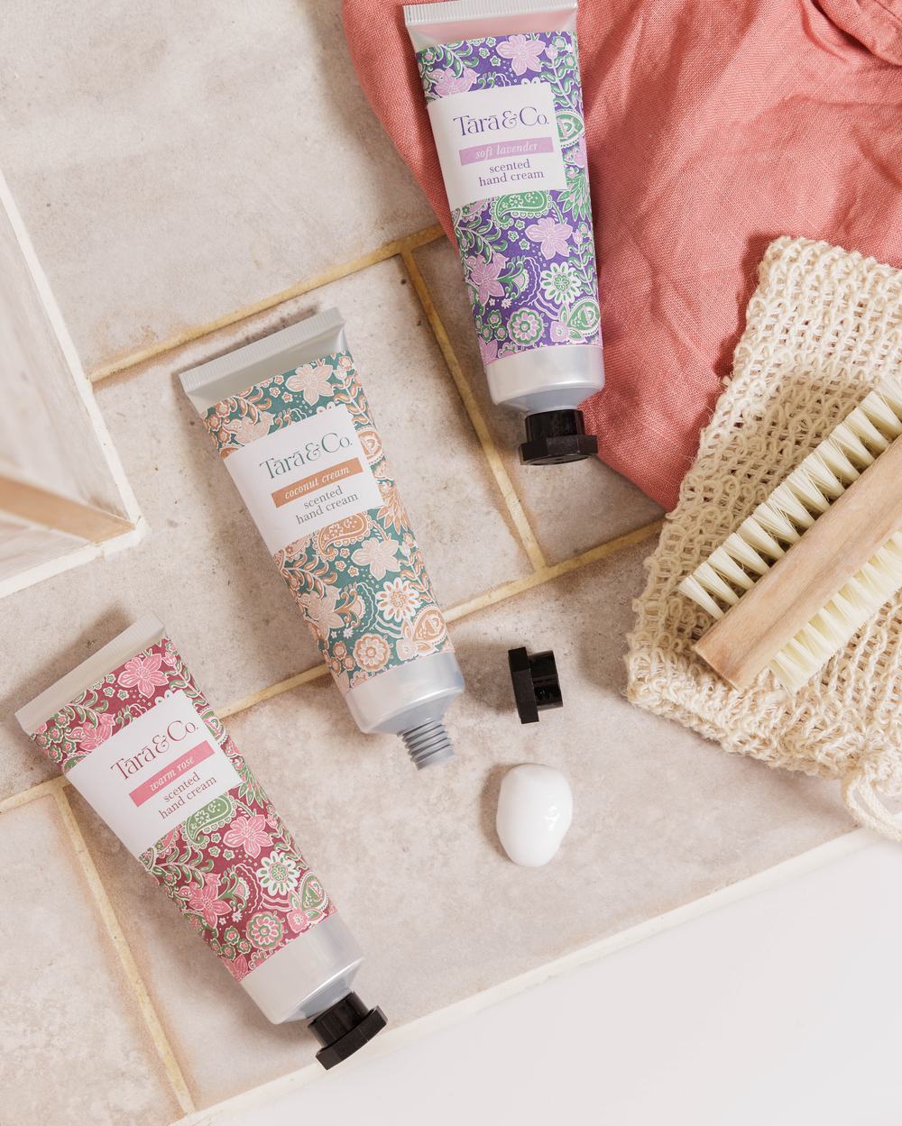 Three tubes of Tara and Co hand cream with floral packaging, laid onto cream tiles.