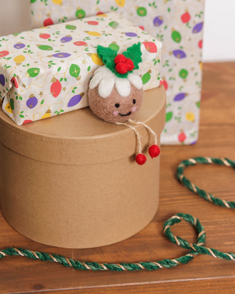 A gift wrapped present and Christmas pudding decoration sit on top of a round cardboard gift box.