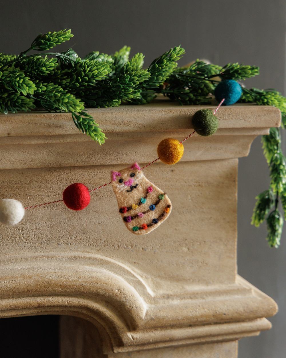 A colourful pompom garland with felted cat decoration hangs from a festive mantelpiece.