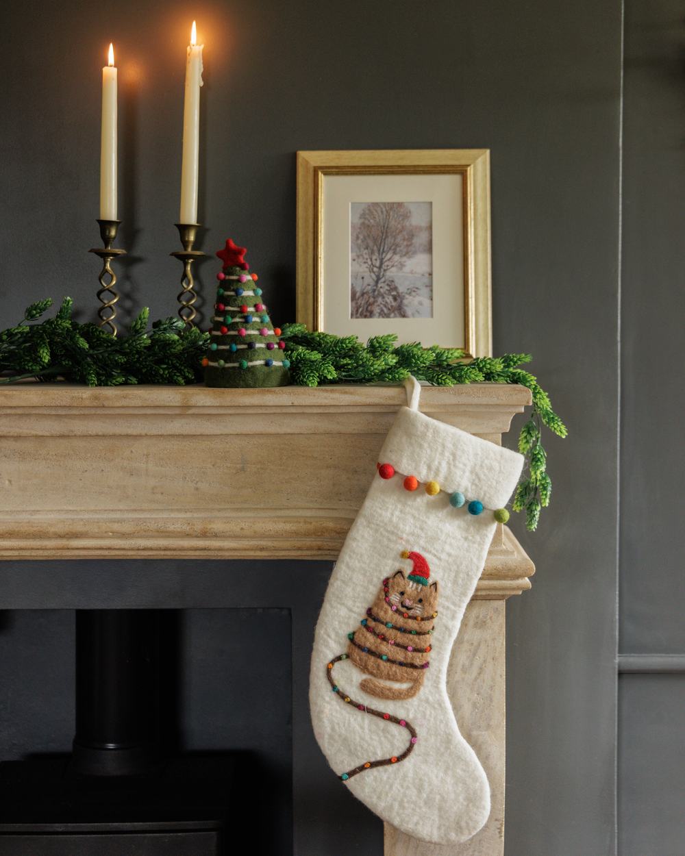 A festive mantelpiece at Christmas, featuring lit candles, a tree ornament, and a felted hanging stocking with a cat design.