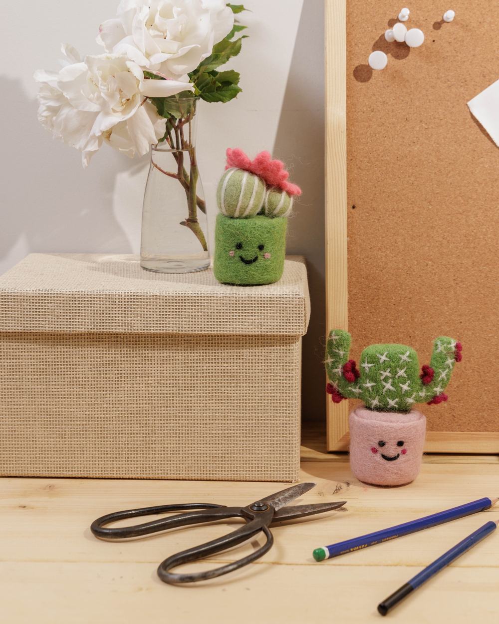 Handcrafted, smiling felt cactus decorations arranged on a desk with flowers, stationery and a corkboard.