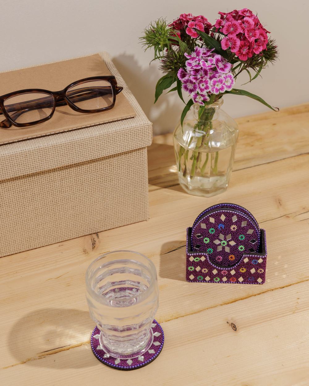 Bejewelled purple mirror and matching coaster on a wooden table, next to a vase of fresh flowers.