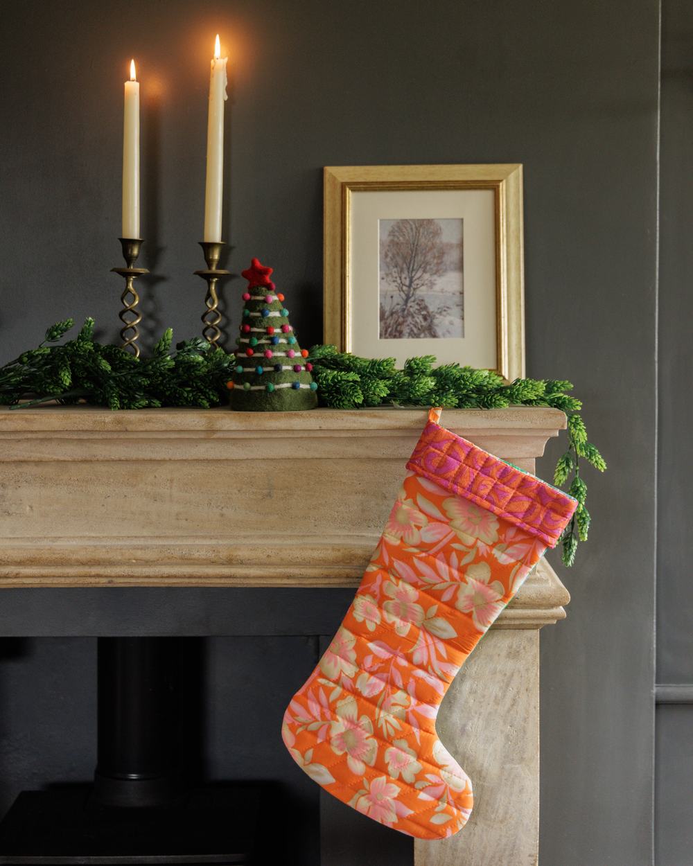A festive mantelpiece at Christmas, featuring lit candles, a tree ornament, and a hanging stocking made from orange floral fabric.