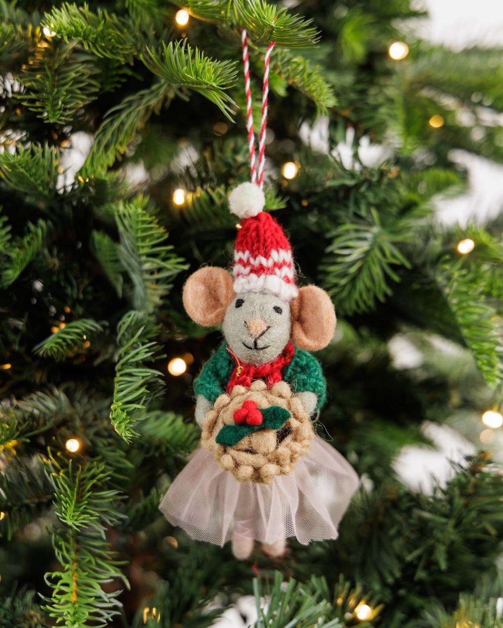 A handmade felt mouse Christmas decoration wearing a red-and-white knitted hat and green cardigan, hanging from a Christmas tree.