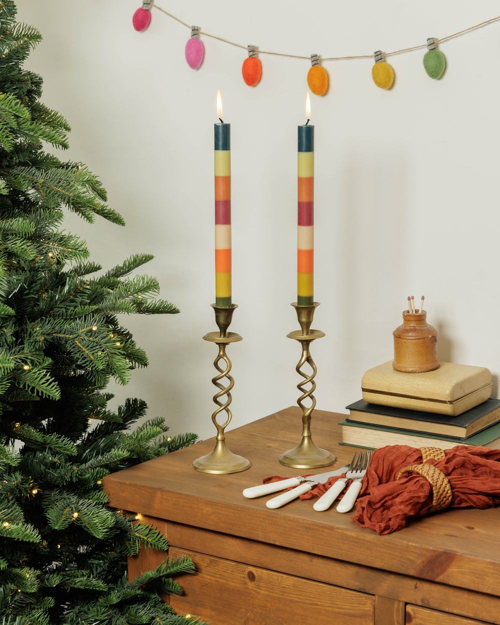 Pair of colourful striped dinner candles in brass holders displayed beside a Christmas tree.