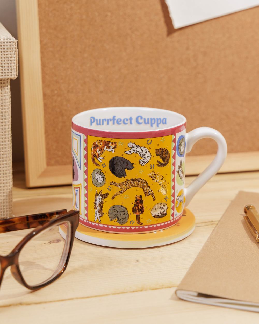 Colourful illustrated mug featuring sleeping cats, displayed on a desk with stationery. The words 'Purrfect Cuppa' are printed on the inside rim.