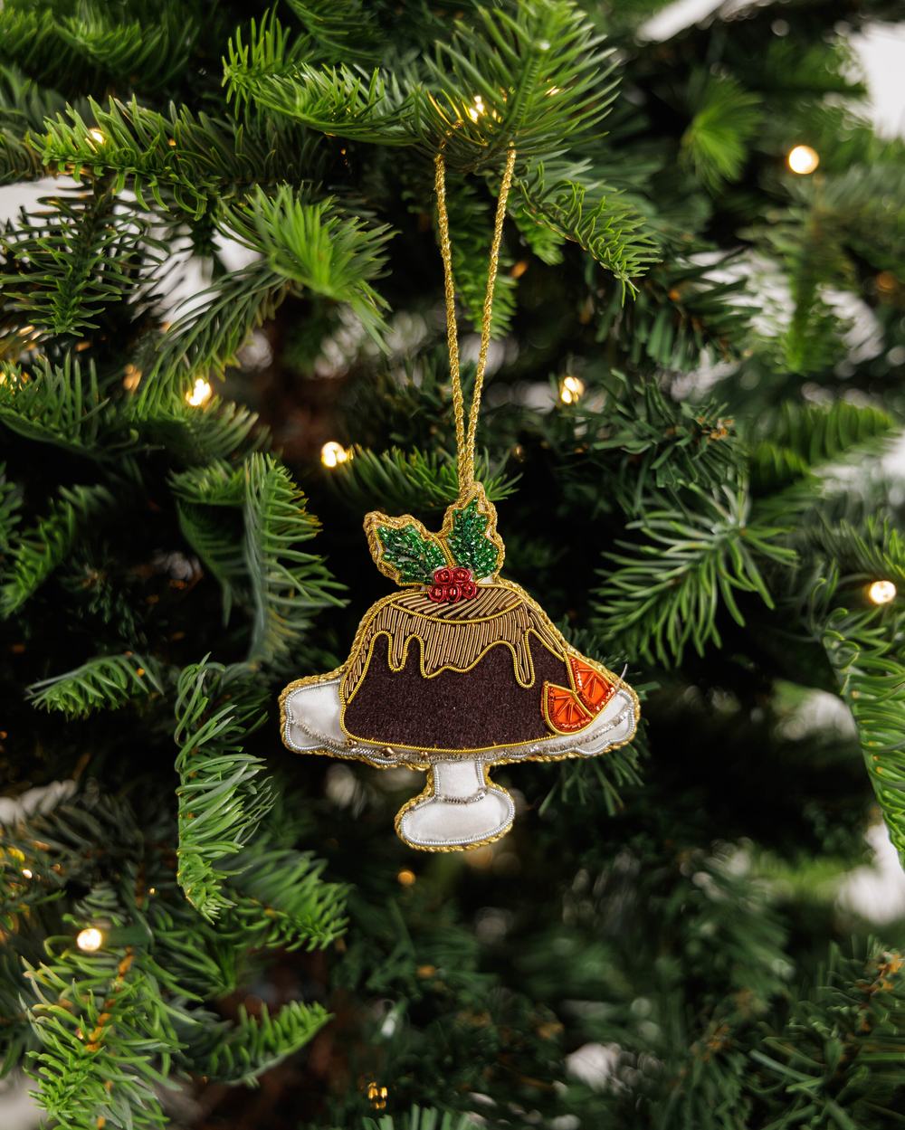 A gold-detailed tree decoration in shape of Christmas pudding, hanging from a Christmas tree lit by fairy lights.