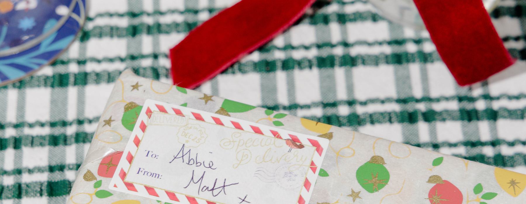 A wrapped Christmas gift with a colourful bauble-patterned paper and handwritten gift tag sits on a green checked festive table setting.