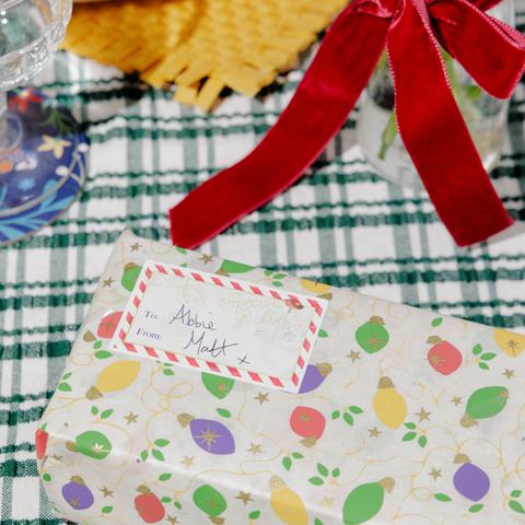 A wrapped Christmas gift with a colourful bauble-patterned paper and handwritten gift tag sits on a green checked festive table setting.