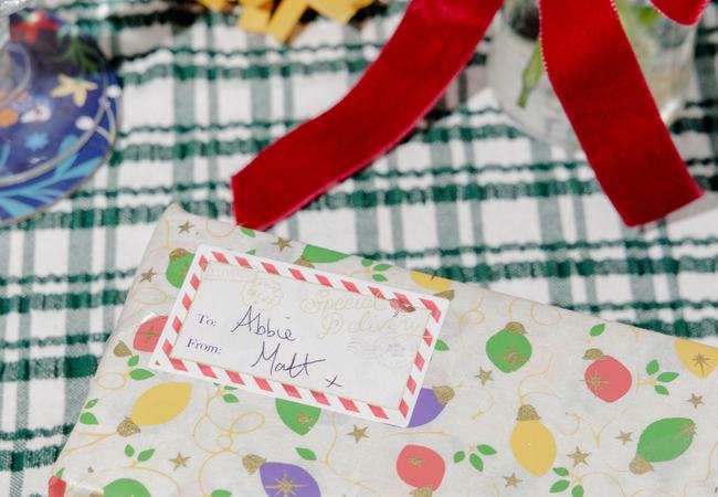 A wrapped Christmas gift with a colourful bauble-patterned paper and handwritten gift tag sits on a green checked festive table setting.