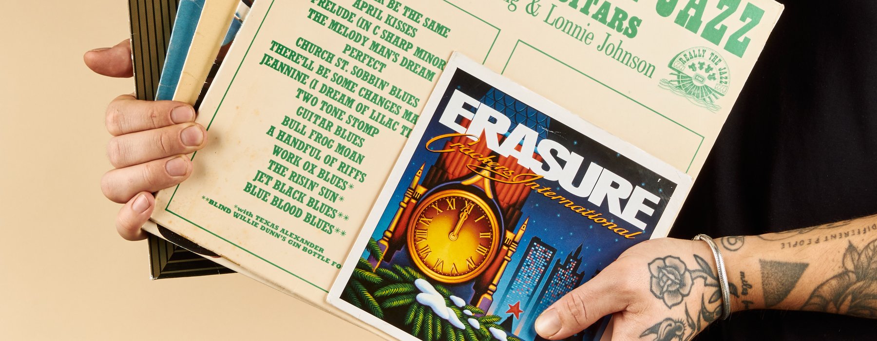 A person with tattoos on their arm holds a stack of vinyl records, with visible covers including "Really the Jazz" and "Erasure."