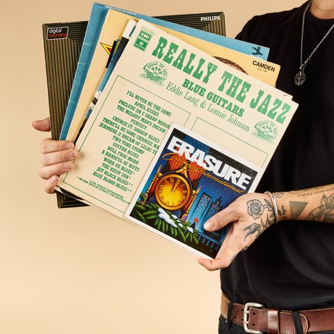 A person with tattoos on their arm holds a stack of vinyl records, with visible covers including "Really the Jazz" and "Erasure."