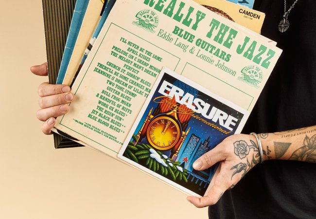 A person with tattoos on their arm holds a stack of vinyl records, with visible covers including "Really the Jazz" and "Erasure."