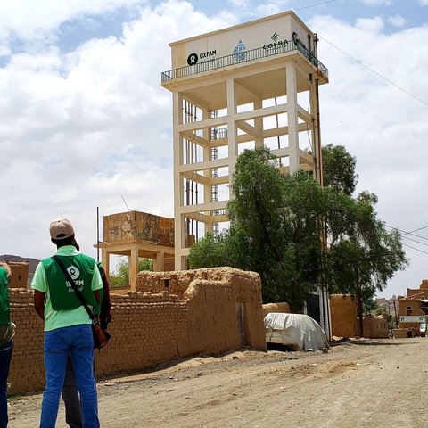 Three people in green Oxfam vests look up at a rainwater harvesting tank rising above the dusty landscape.