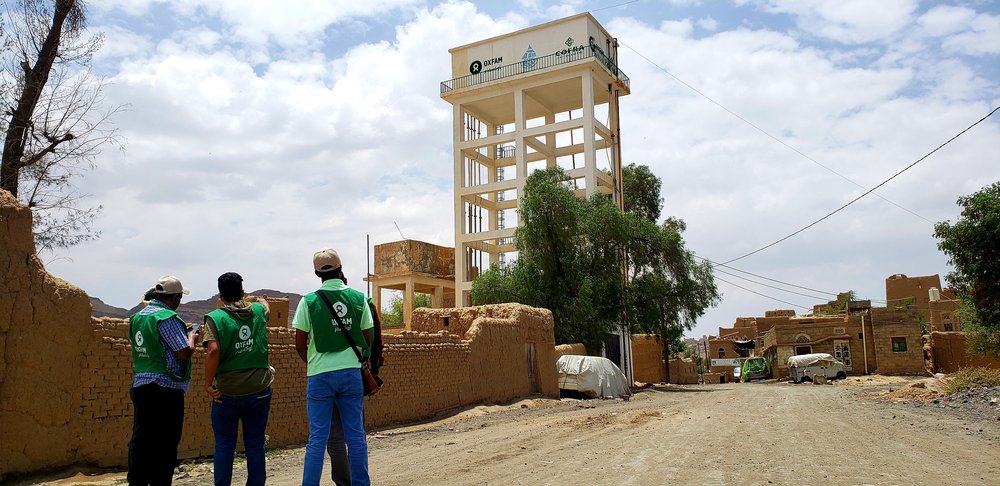 Three people in green Oxfam vests look up at a rainwater harvesting tank rising above the dusty landscape.