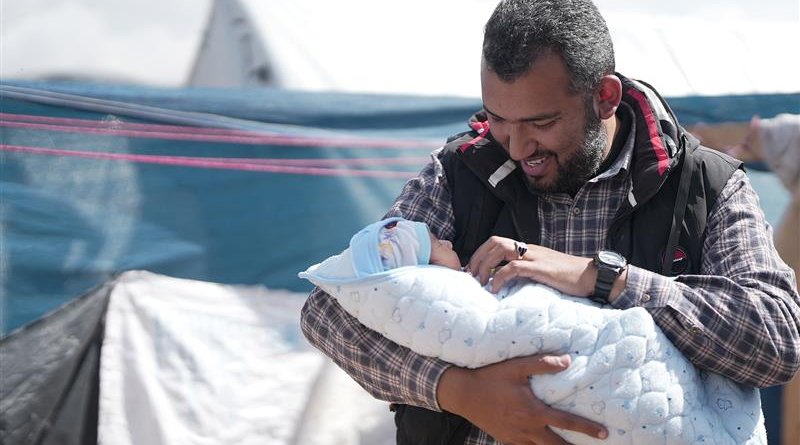 Fedaa  is smiling warmly while holding a newborn baby wrapped in a light blue blanket with a subtle pattern. The baby is also wearing a small blue cap. The background consists of a refugee or