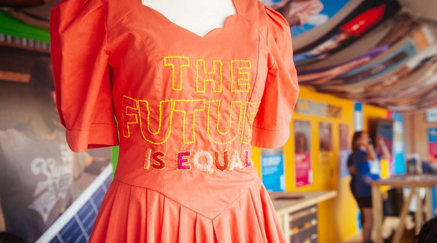 An orange dress on a mannequin with the stitched slogan 'The future is equal'.