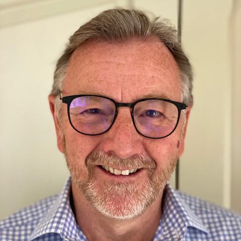 A smiling man with glasses, grey hair and a beard, wearing a checked shirt.