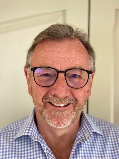 A smiling man with glasses, grey hair and a beard, wearing a checked shirt.