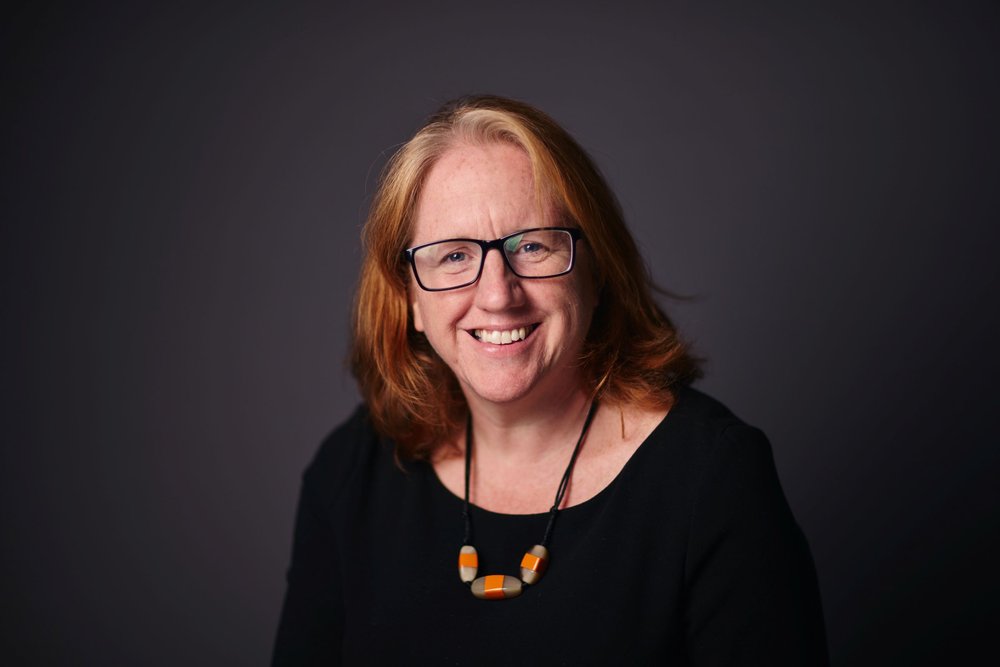 A woman with shoulder-length red hair and black glasses smiles into the camera. She is wearing a black top and an orange necklace. The background is a plain dark grey.