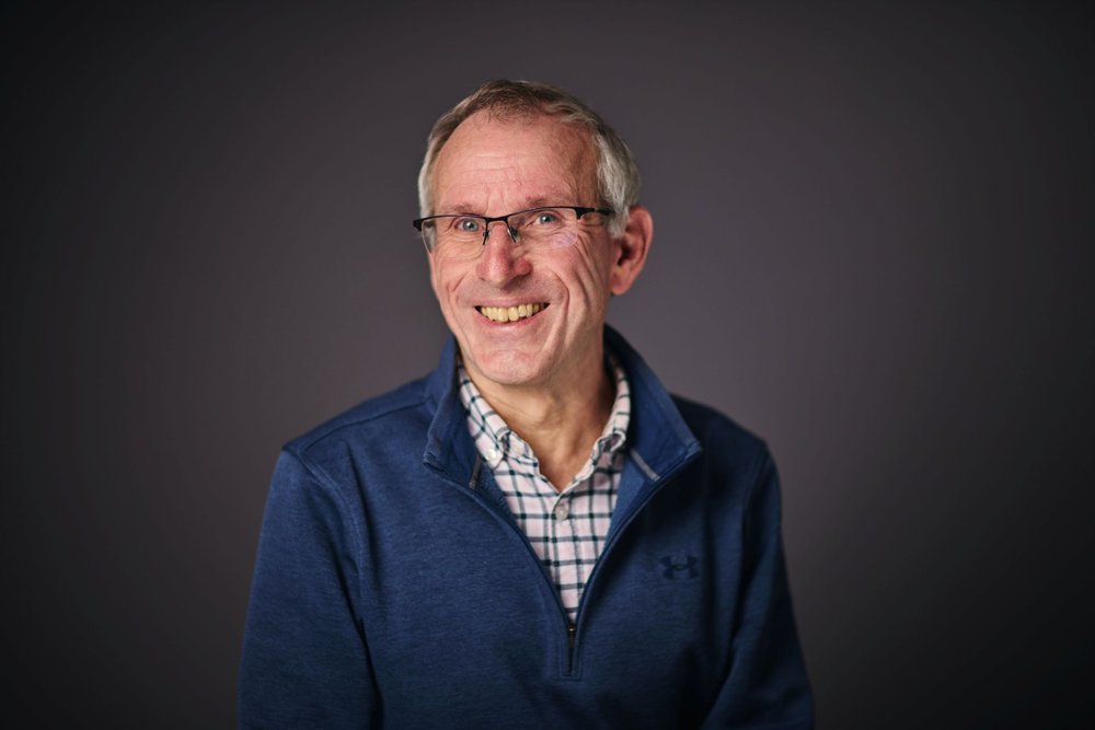 An older man wearing glasses, a checked shirt and a dark blue zip-up jumper smiles into the camera, posed against a dark background.