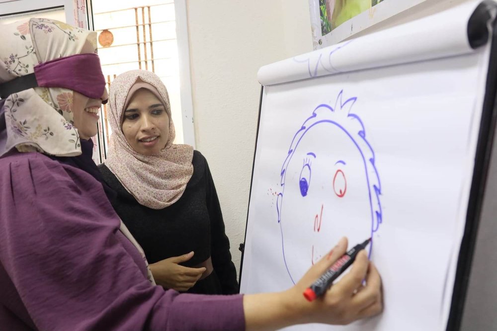 Two women, one blindfolded are smiling and drawing a picture of a face on a flip chart.
