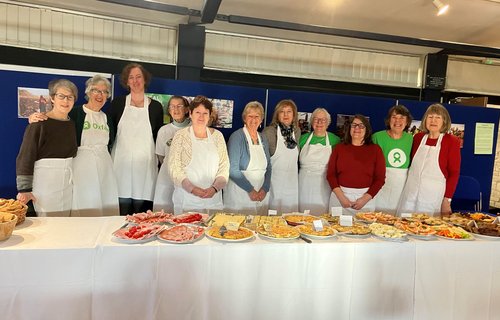 A group of people wearing white aprons stand behind a long table covered with various plates of food, including pastries, bread, and sliced meats, in an indoor event setting.