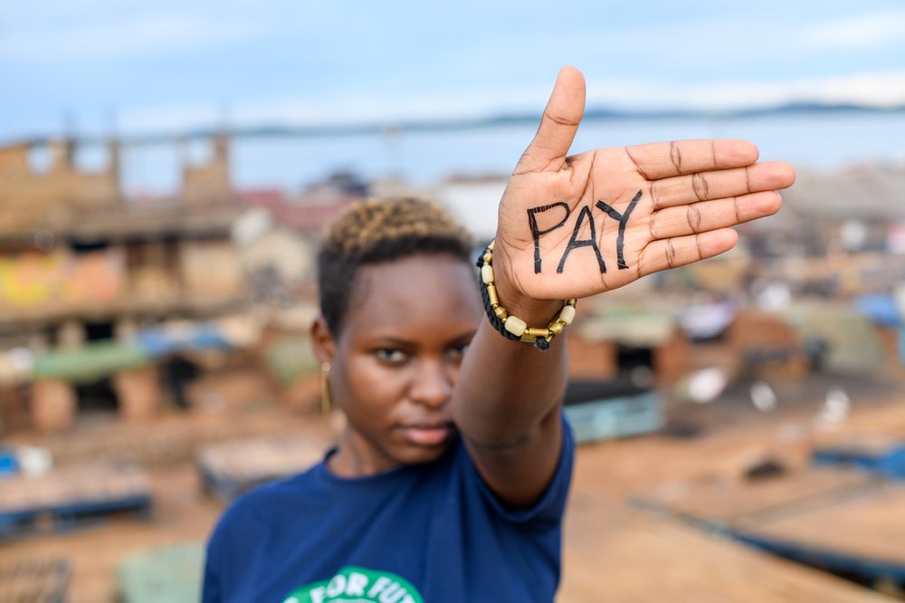 A woman holds out her hand confidently, with the word "PAY" inked across her palm. In the background, coastal buildings line the horizon.