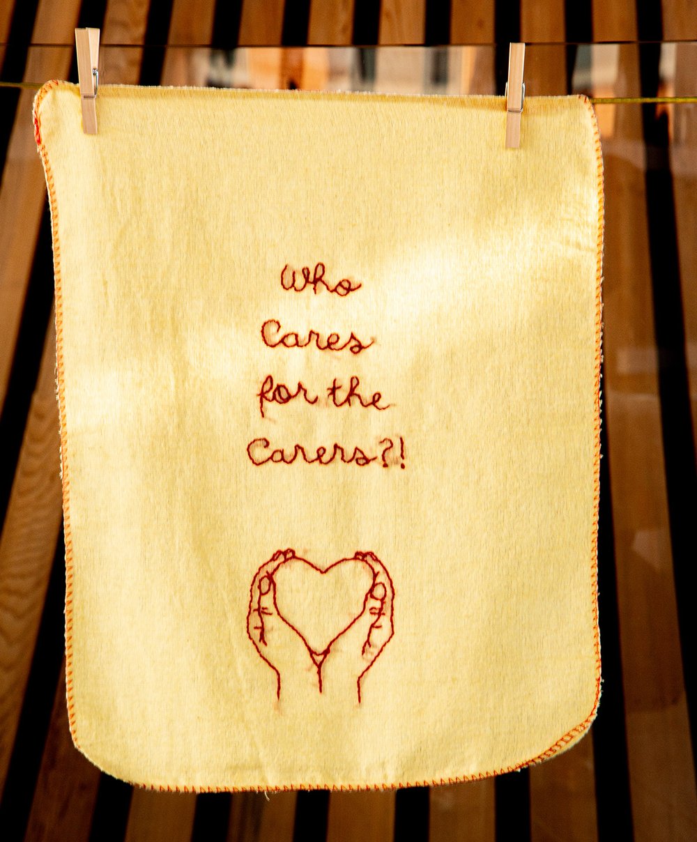 An embroidery with two hands holding a heart and the text 'Who cares for carers?'